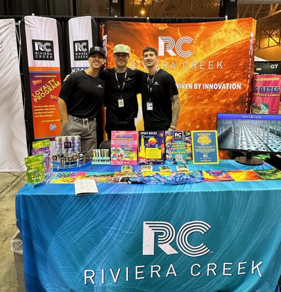 Three Riviera Creek team members standing behind a branded booth at a trade show, displaying RC products, promotional materials, and signage that reads “Driven by Innovation.”