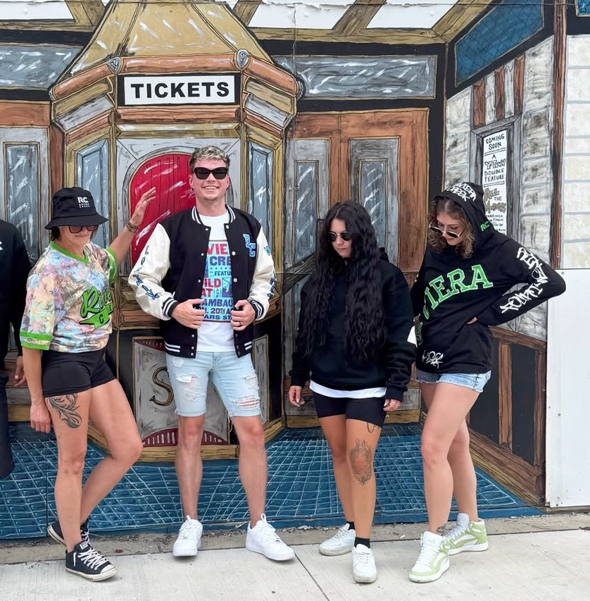 Four people posing in front of a vintage-style “Tickets” mural, wearing Riviera Creek branded apparel including hoodies, hats, and jackets.
